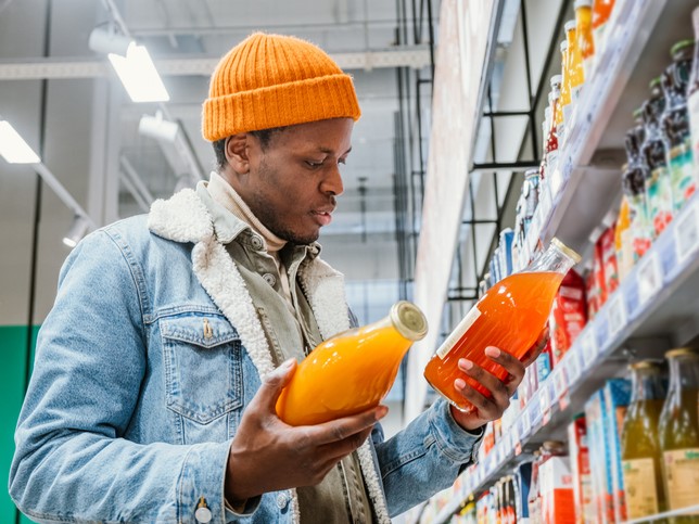 Afrikansk mann med en stilig oransje hatt og olajakke velger naturlig juice i glassflasker i et dagligvaremarked